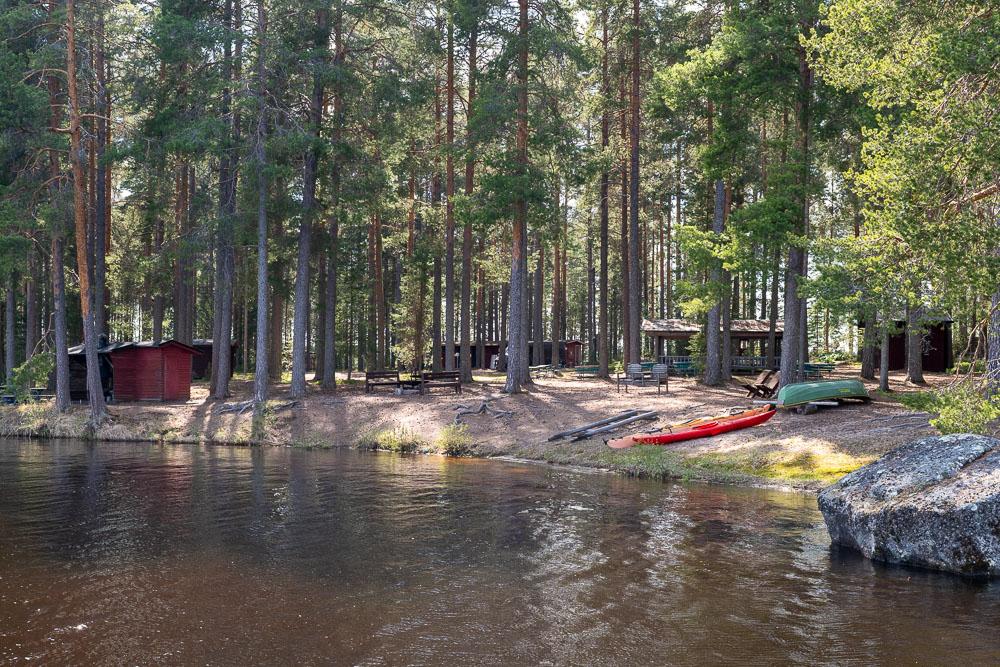 Camping Sumiainen overview of the shore. Trees, a rowboat and cottages by the water.